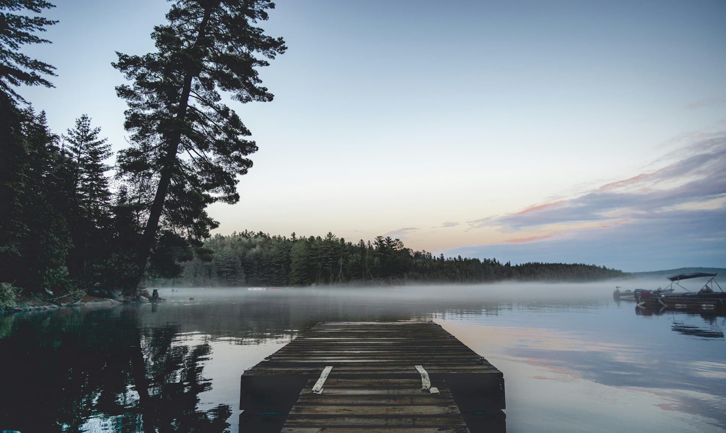 A weathered wooden dock running out into a still lake at first light, mist on the water and white pines along the shore.